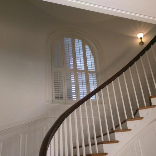 White plantation shutters adorning arched window located in curved stairwell.